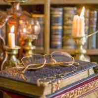 A cozy home library scene with a collection of antique biographies, a lit candle, and a pair of eyeglasses resting on top of an old book.