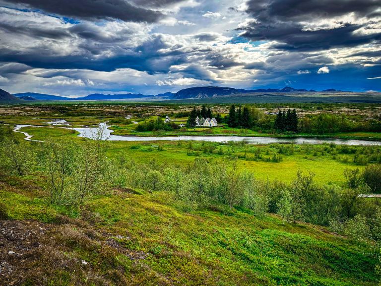 Dramatic storm clouds over a green Icelandic valley with a winding river, distant mountains, and small white houses