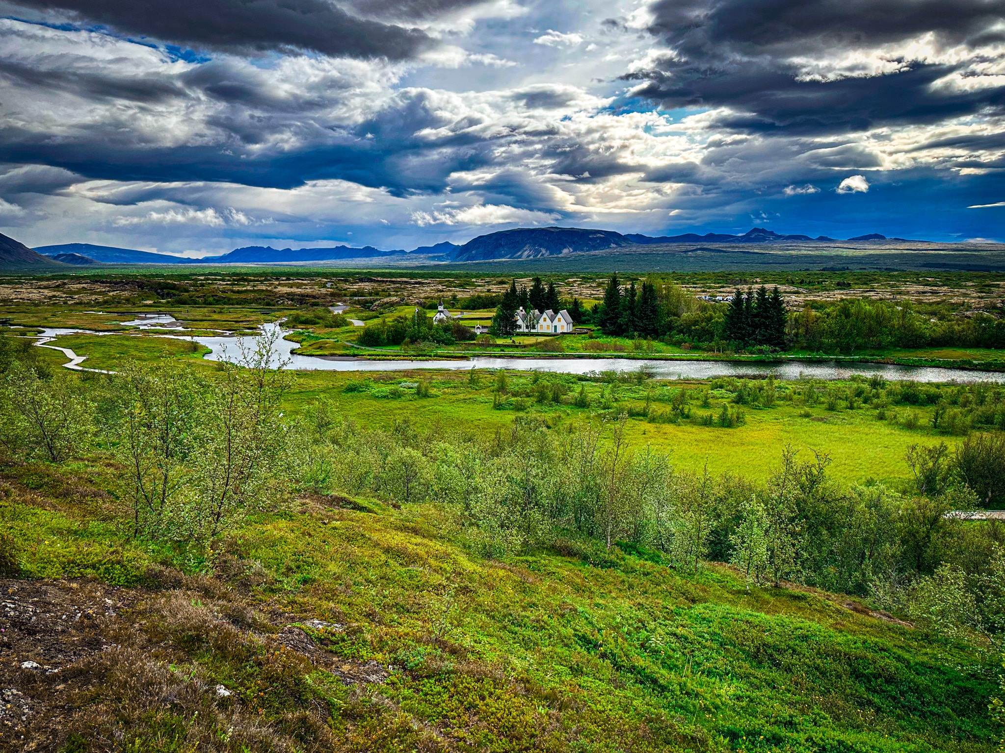 Dramatic storm clouds over a green Icelandic valley with a winding river, distant mountains, and small white houses