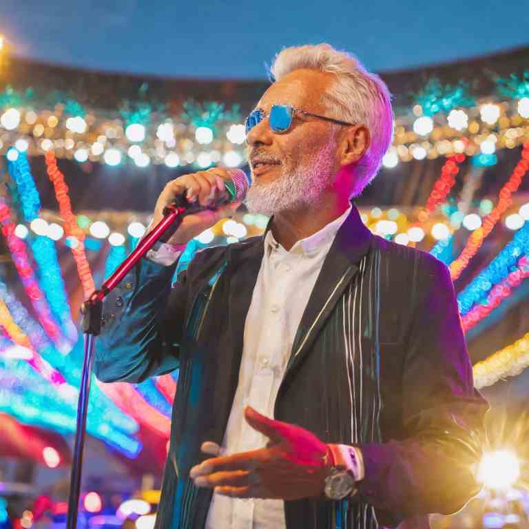 A senior man with silver hair, sporting blue sunglasses and a chic black jacket, confidently sings into a microphone on a stage aglow with vibrant bokeh lights.