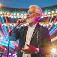A senior man with silver hair, sporting blue sunglasses and a chic black jacket, confidently sings into a microphone on a stage aglow with vibrant bokeh lights.