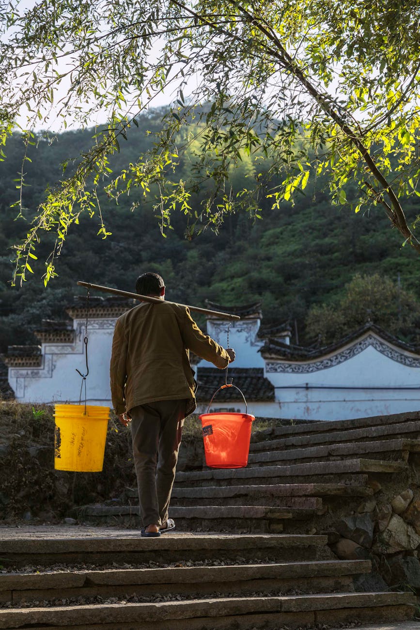 anonymous man carrying buckets with water using shoulder yoke