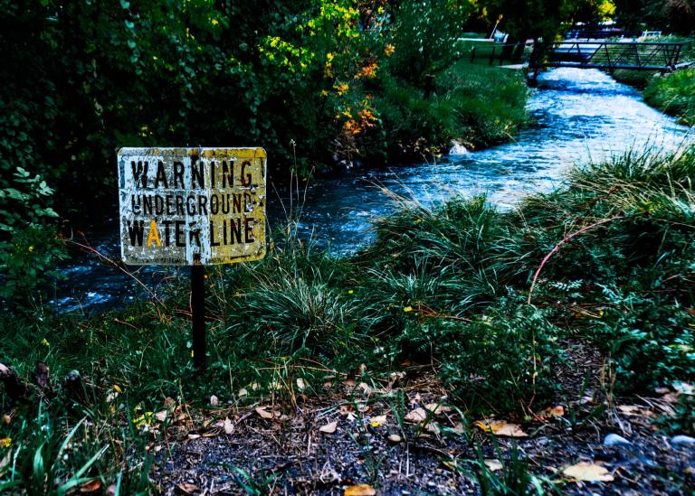 A shallow Utah creek flowing beneath trees beside a weathered underground water line warning sign.